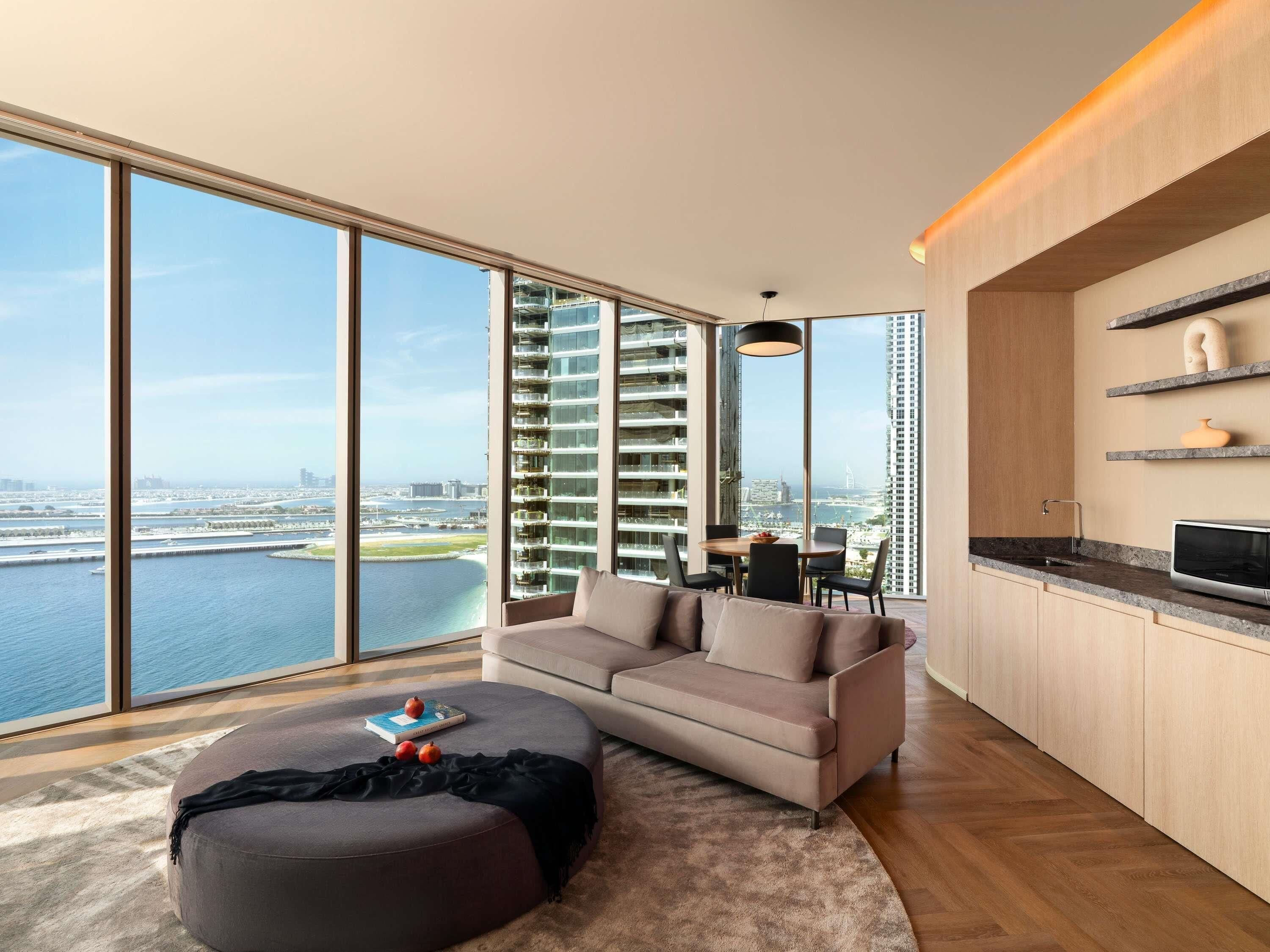 A modern living room with a large window, a sofa, a dining table, and a kitchen area.
