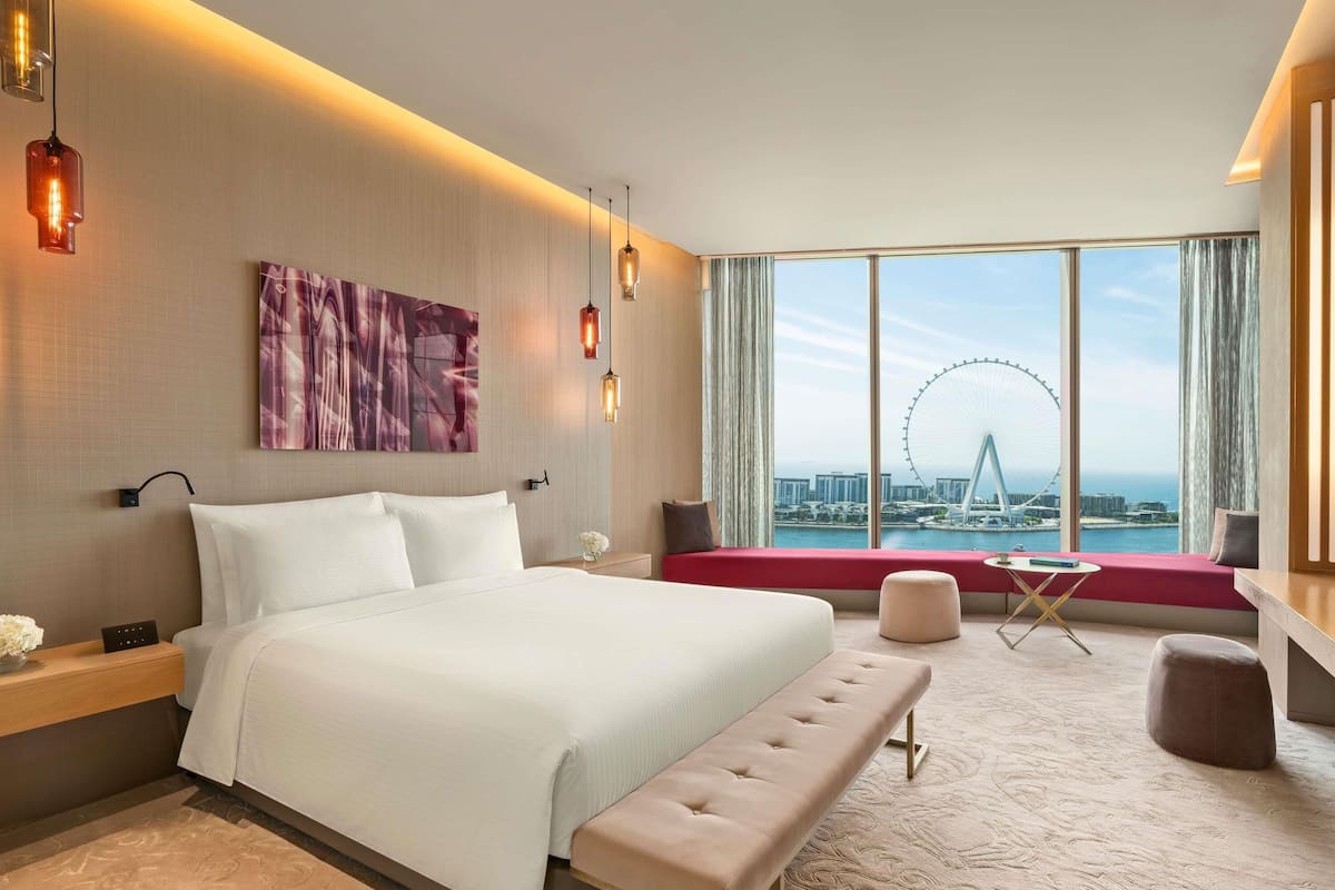 A modern hotel room with a large bed, a bench, a view of a Ferris wheel, and a cityscape.