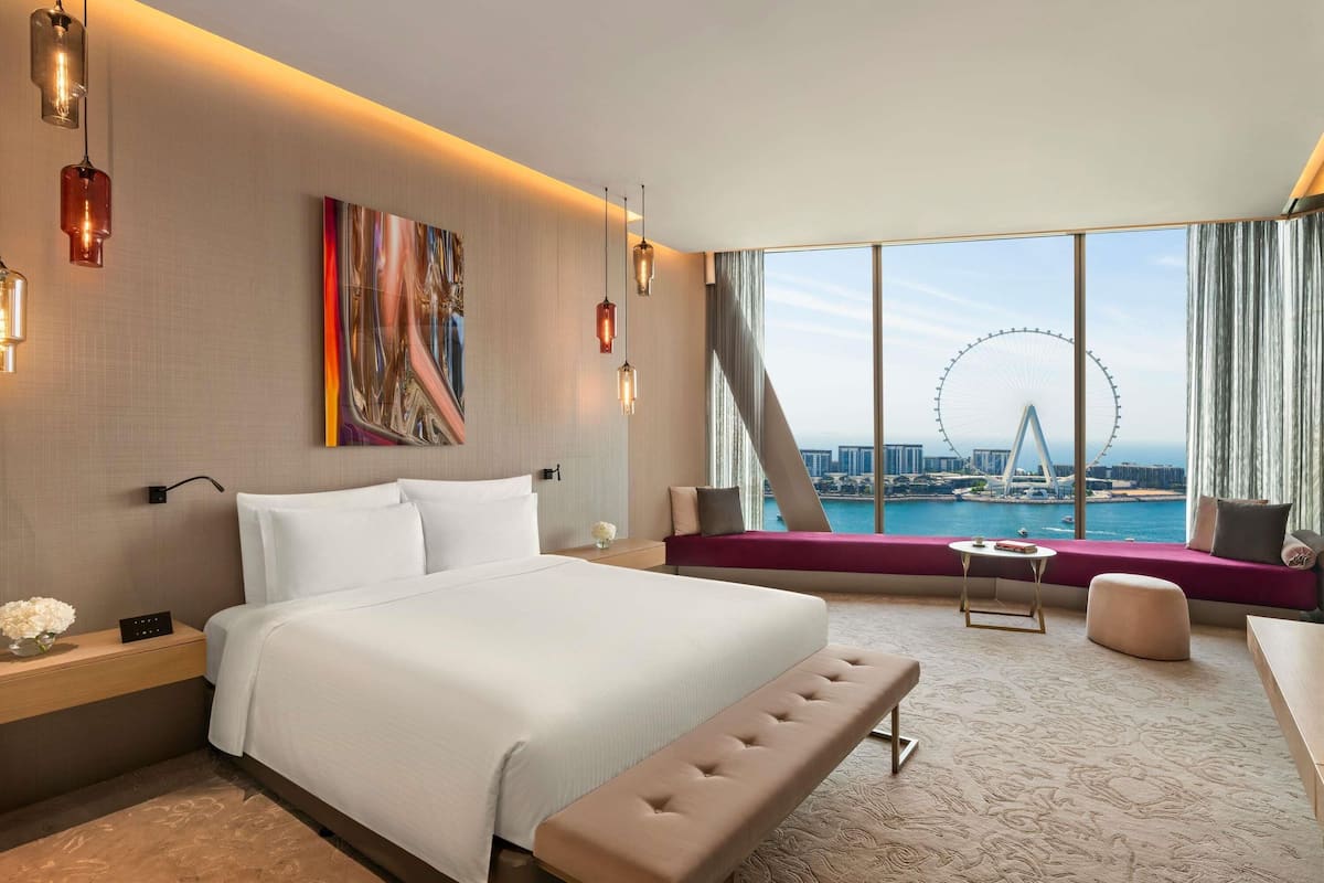 A modern hotel room with a large bed, a bench, a small table, and a view of a Ferris wheel and bridge.