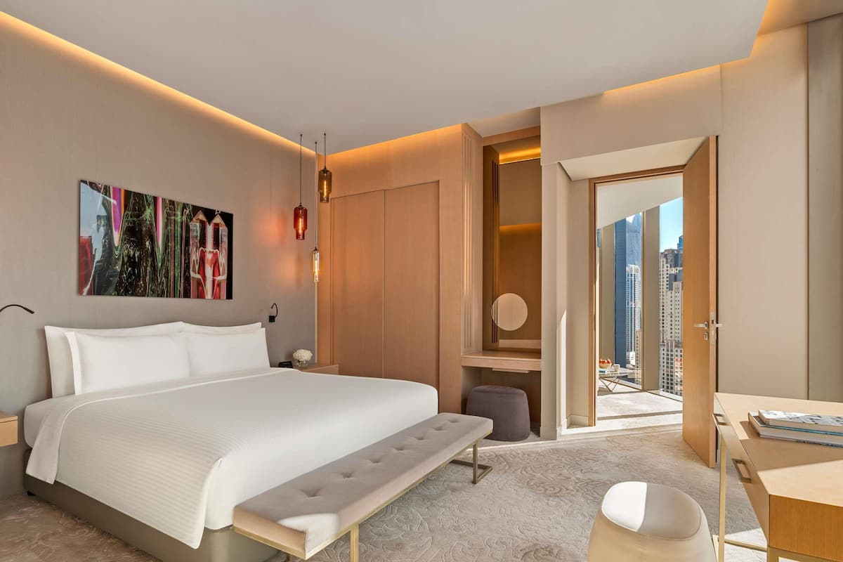 A modern hotel room with a bed, bedside table, bench, desk, and a view of the city.