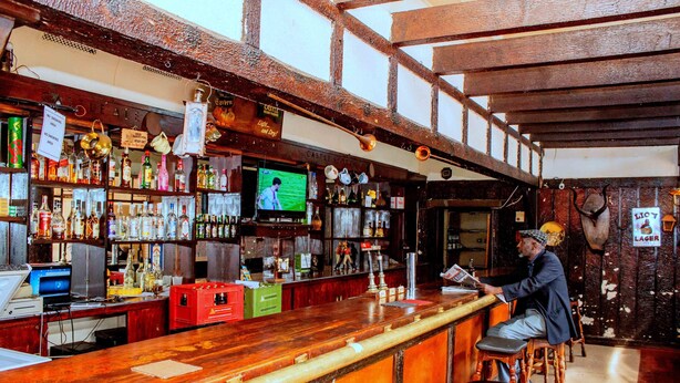 A bar with wooden beams, shelves of bottles, and a television screen. - The Grey's Inn (Bulawayo)
