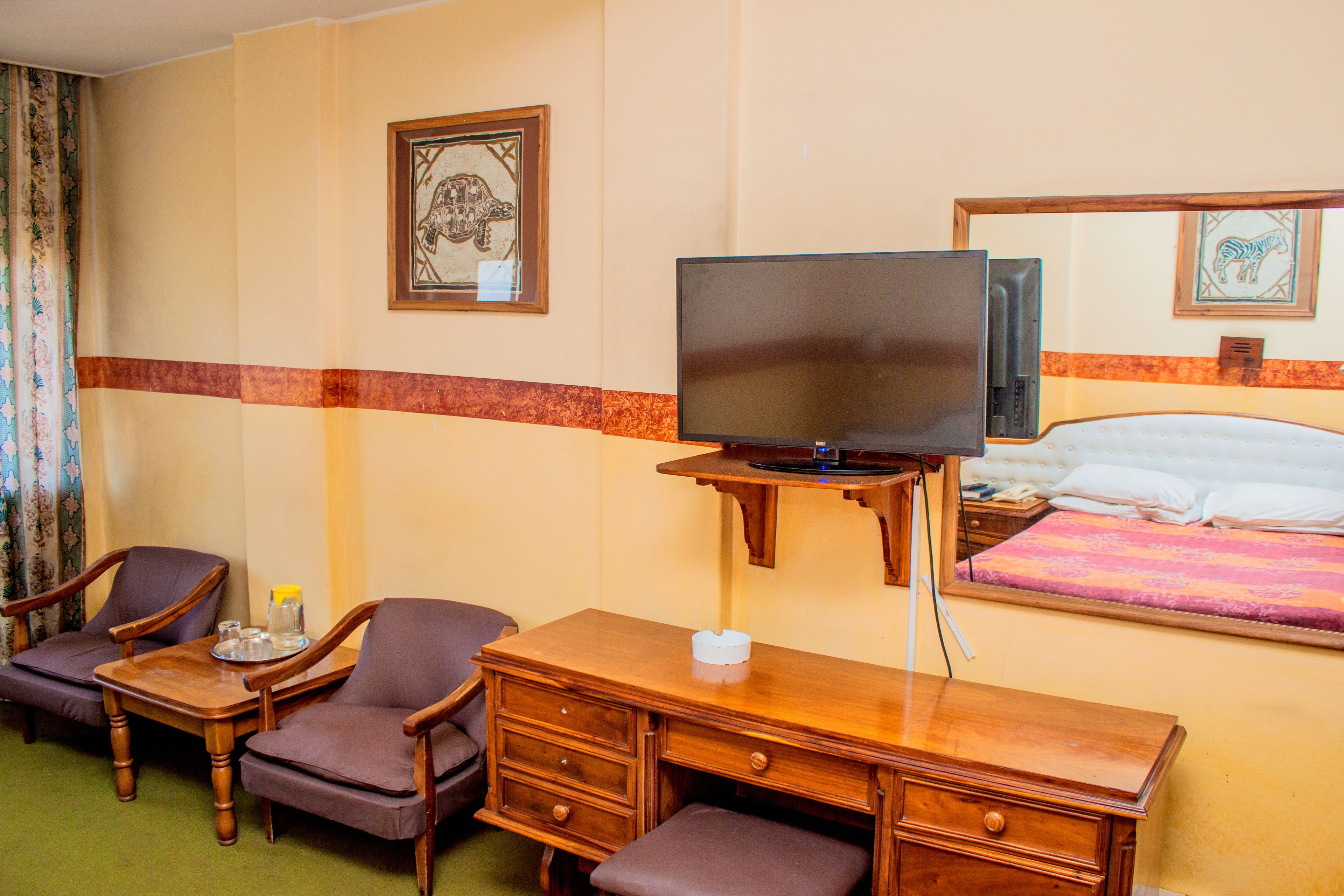 A hotel room with a bed, a television, a desk with drawers, two armchairs, and a small table with a glass and a bottle.