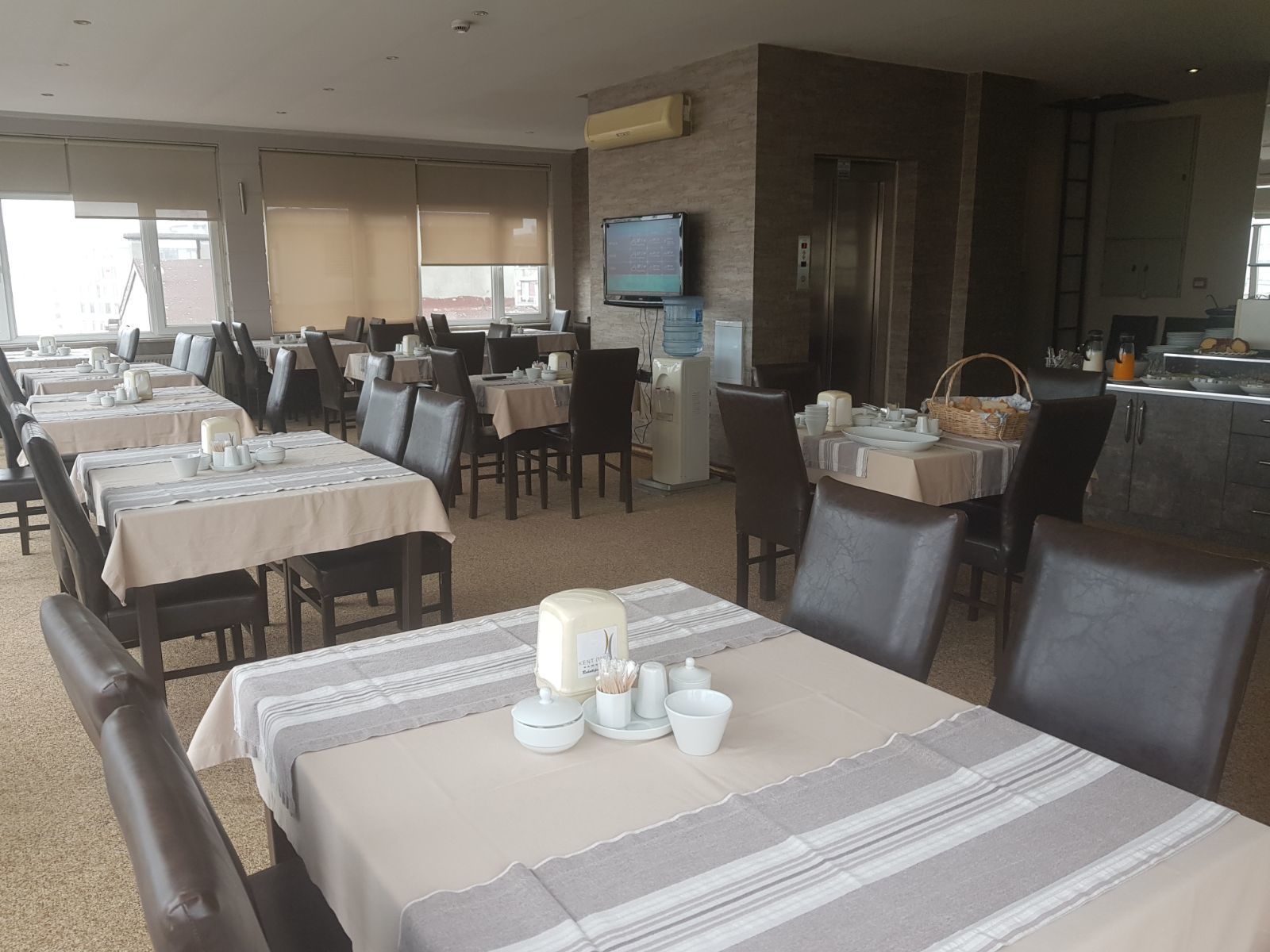 Comedor de hotel con varias mesas dispuestas para comer, área de cocina con refrigerador y lavaplatos, y un televisor montado en la pared.