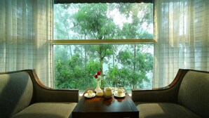Una habitación con un ventanal grande, un sofá y una mesa con tetera, tazas y un florero con una flor.
