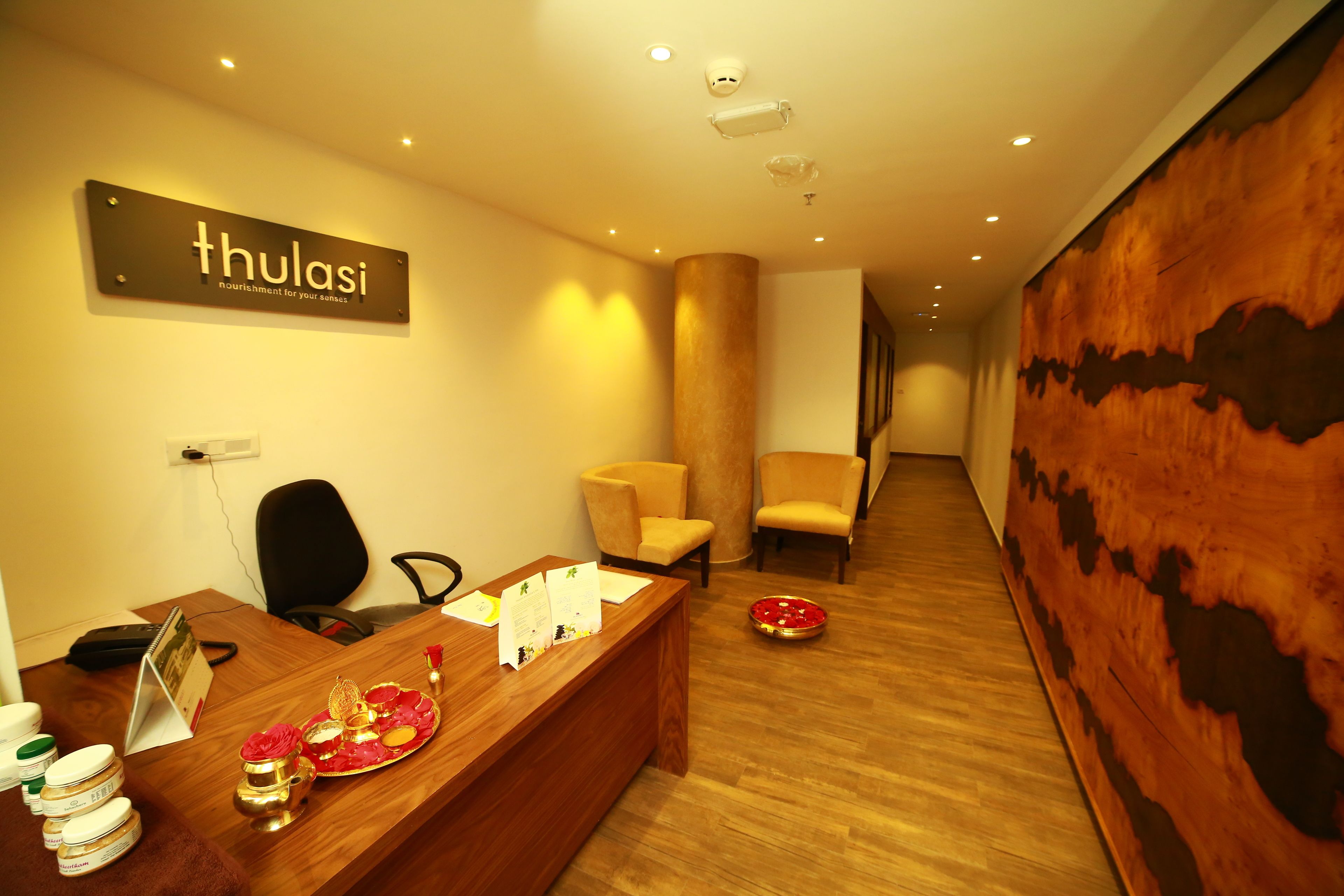 A reception area with a wooden desk, a sign reading "thulasi," and a reception counter. There are two chairs, a wooden wall with a natural pattern, and a wooden floor.