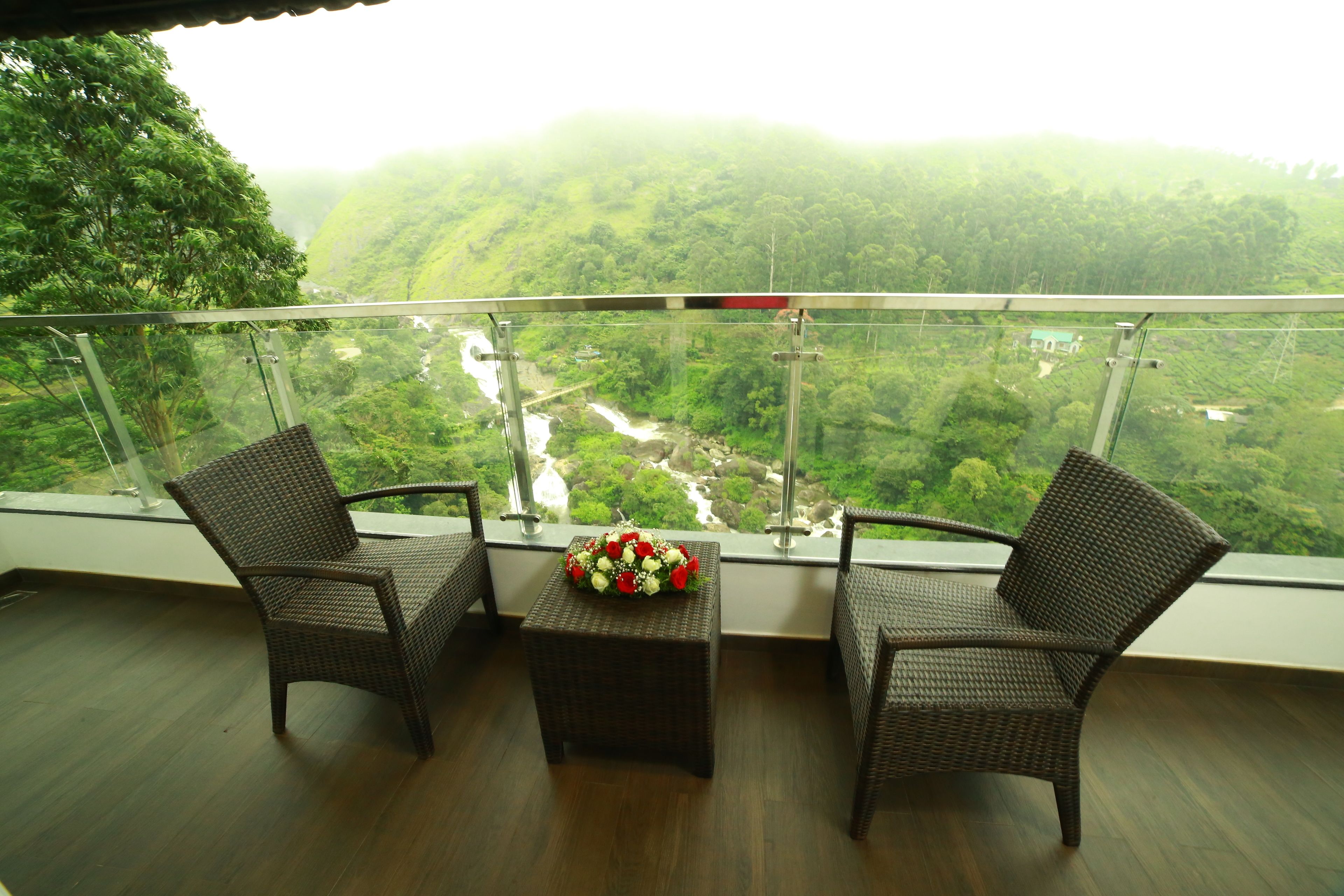 Un balcón con mobiliario de mimbre y un centro de mesa floral, con vistas a un valle exuberante y un río.
