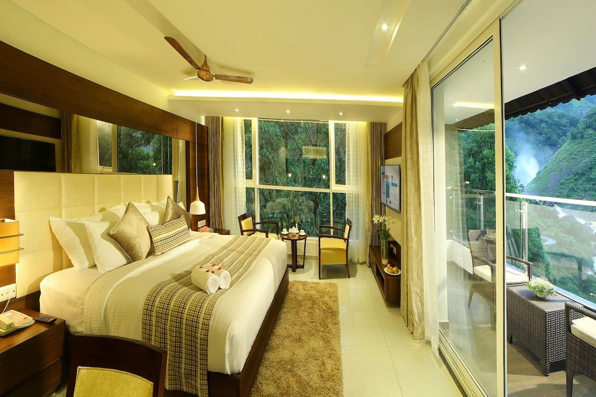 A modern hotel room with a large bed, a desk, a chair, a small table, and a balcony with a scenic view of a waterfall.