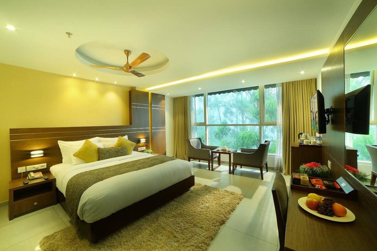 A modern hotel room with a large bed, a ceiling fan, a TV, and a dining area with a fruit plate.