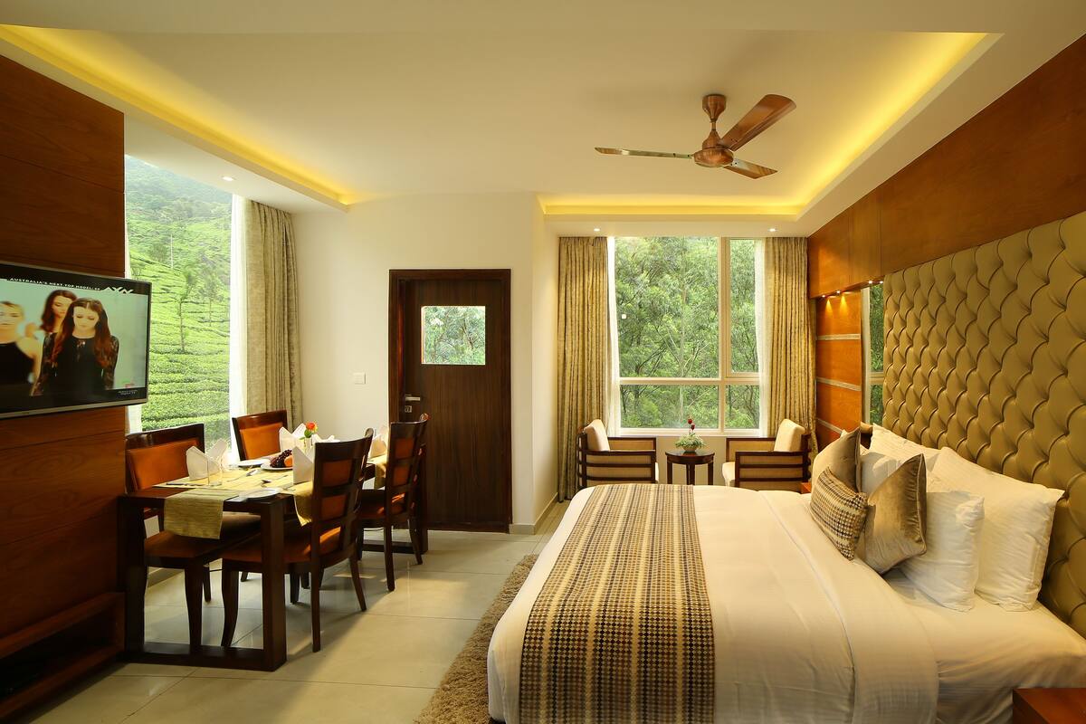 A hotel room with a large bed, a desk, and a chair, a ceiling fan, and a window with a view of greenery.