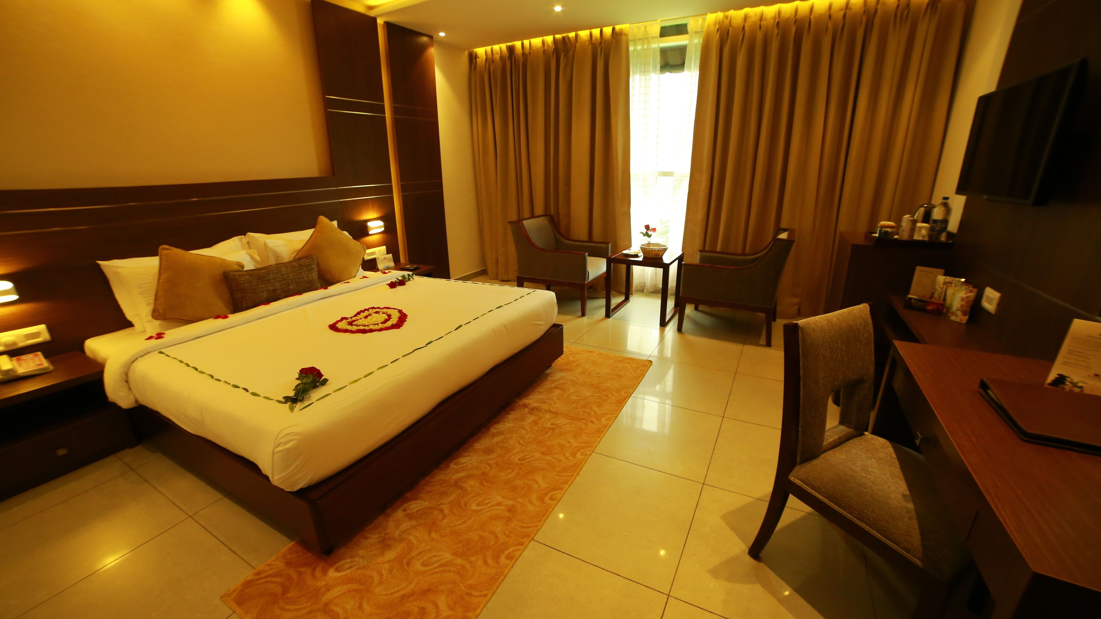 A hotel room with a large bed, a desk, two chairs, a TV, and a nightstand with a lamp.
