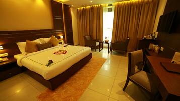 A hotel room with a large bed, a desk, two chairs, a TV, and a nightstand with a lamp.