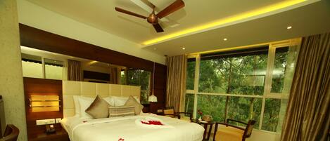 A spacious bedroom with a large bed, a ceiling fan, a bedside table with a lamp, a window with curtains, and a view of greenery outside.