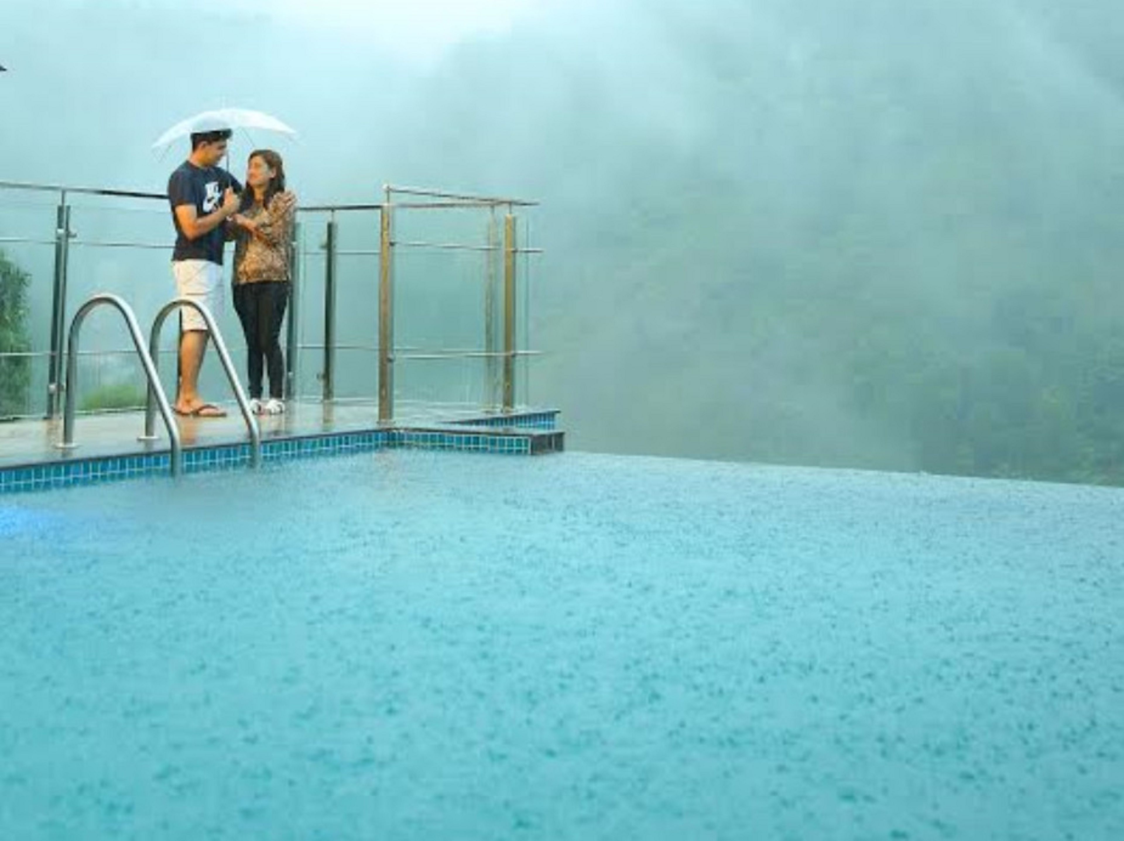 A couple standing by a pool with a glass railing, under a cloudy sky.