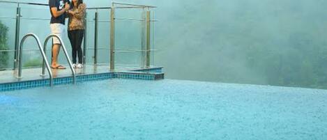 A couple standing by a pool with a glass railing, under a cloudy sky.