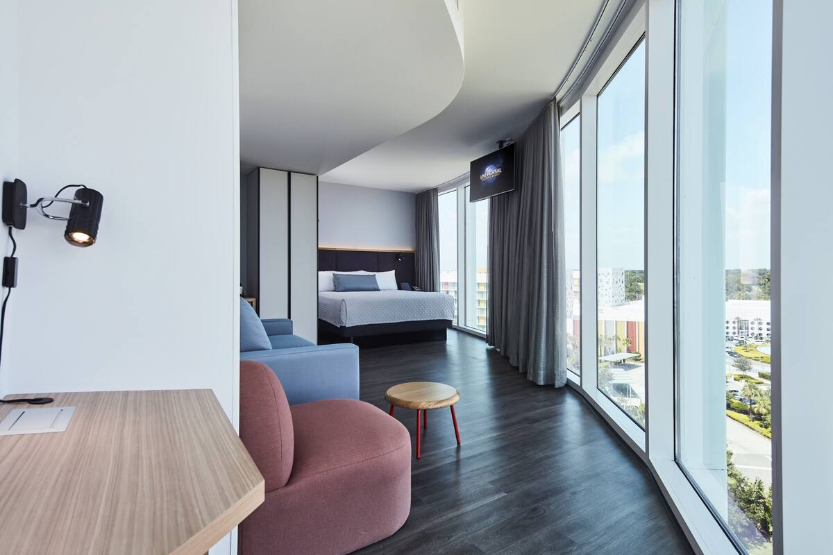 A modern hotel room with a large window, a bed, a desk, and a chair.