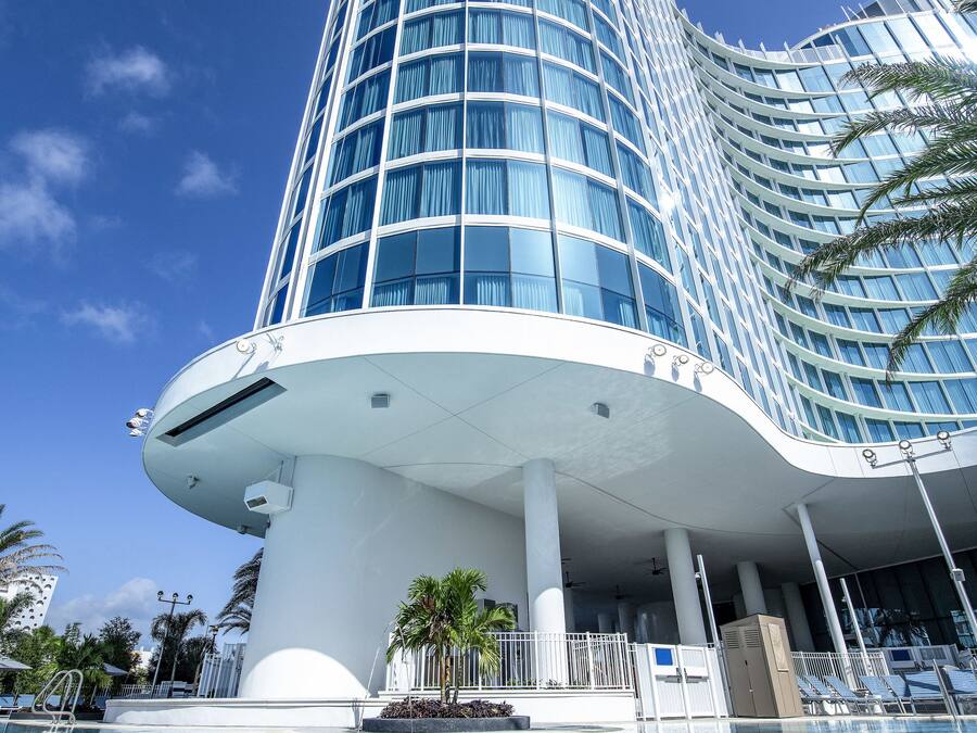 Un hotel moderno con fachada curva, una piscina en primer plano y un cielo azul despejado.