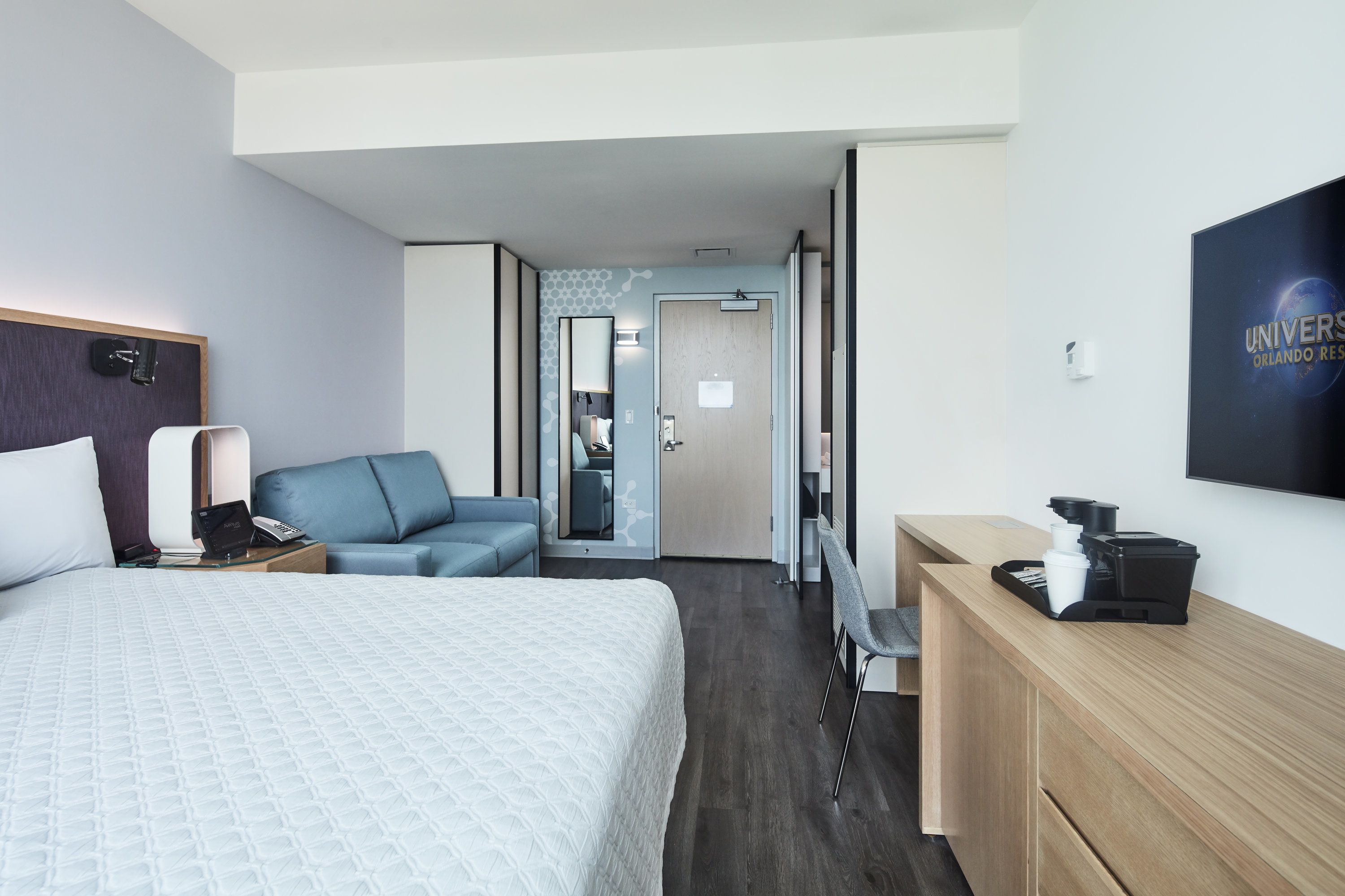 Habitación de hotel con cama, escritorio con cafetera, zona de estar y televisor montado en la pared.