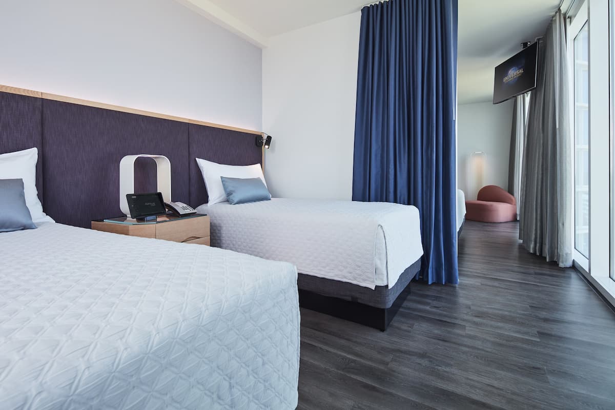 A hotel room with two beds, a desk with a laptop, a flat-screen TV, and a large window with curtains.
