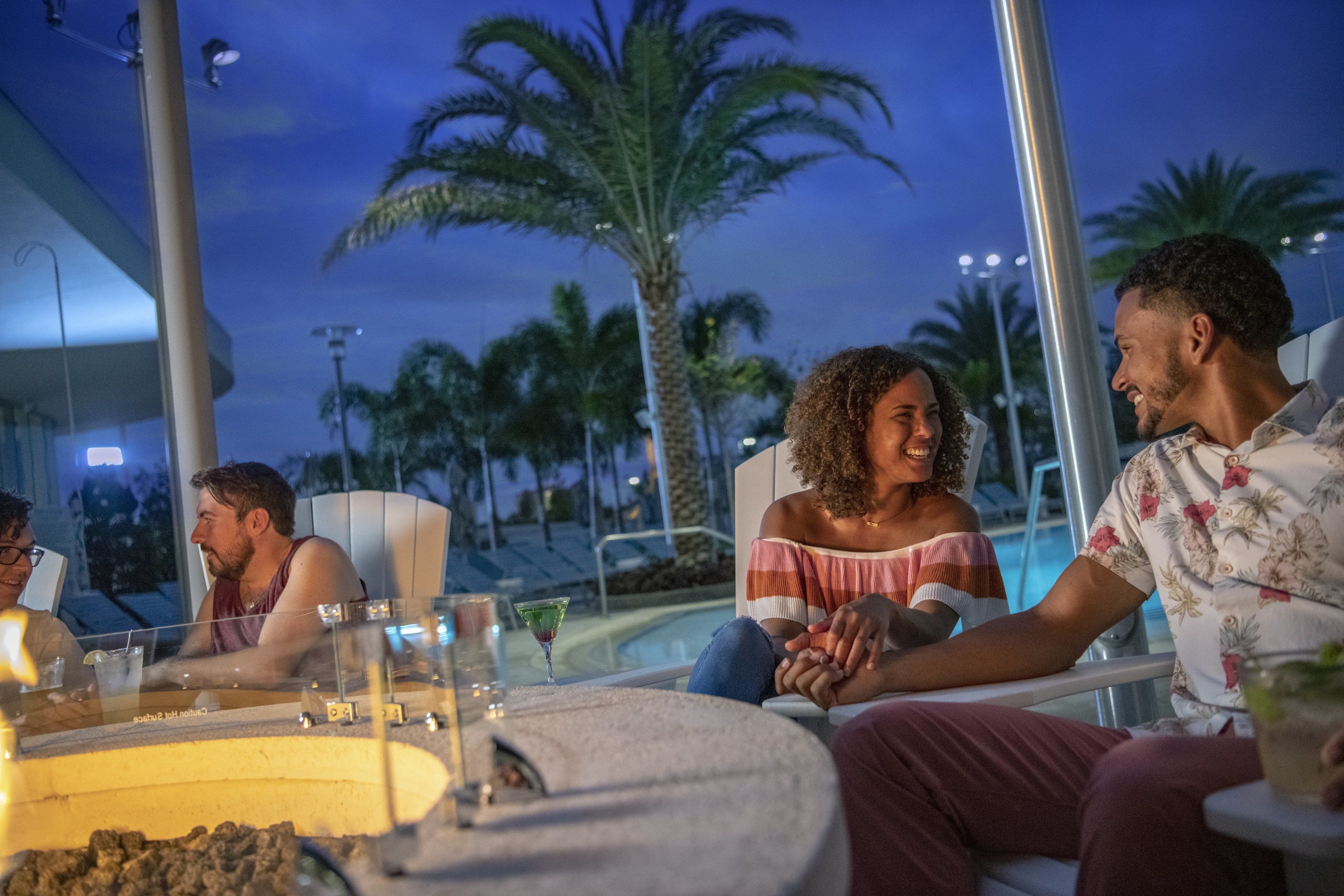 A group of people sitting around a fire pit at an outdoor lounge area with a pool and palm trees in the background.