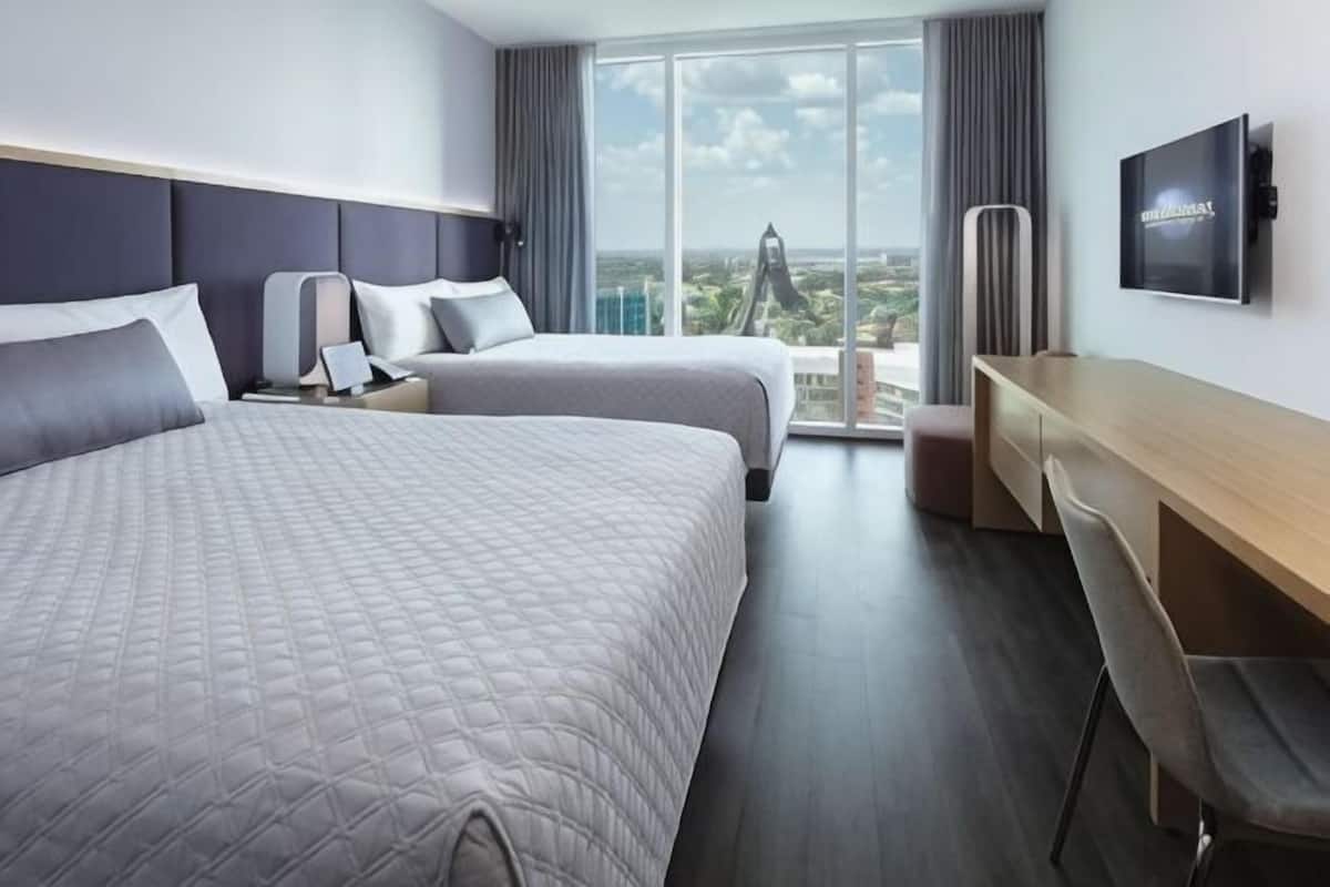 A hotel room with two beds, a desk, a chair, a TV, and a large window with a city view.