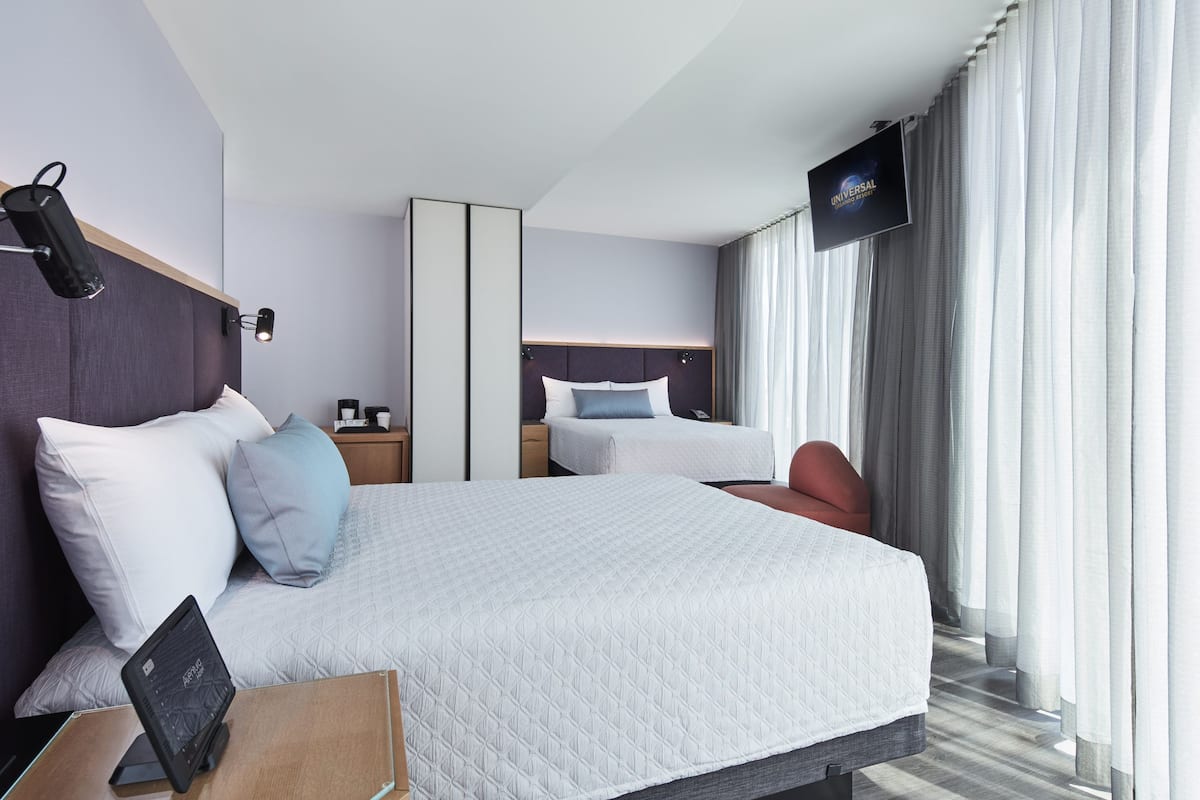 A modern hotel room with a large bed, bedside tables, a flat-screen TV, and a view of the outdoors through large windows.