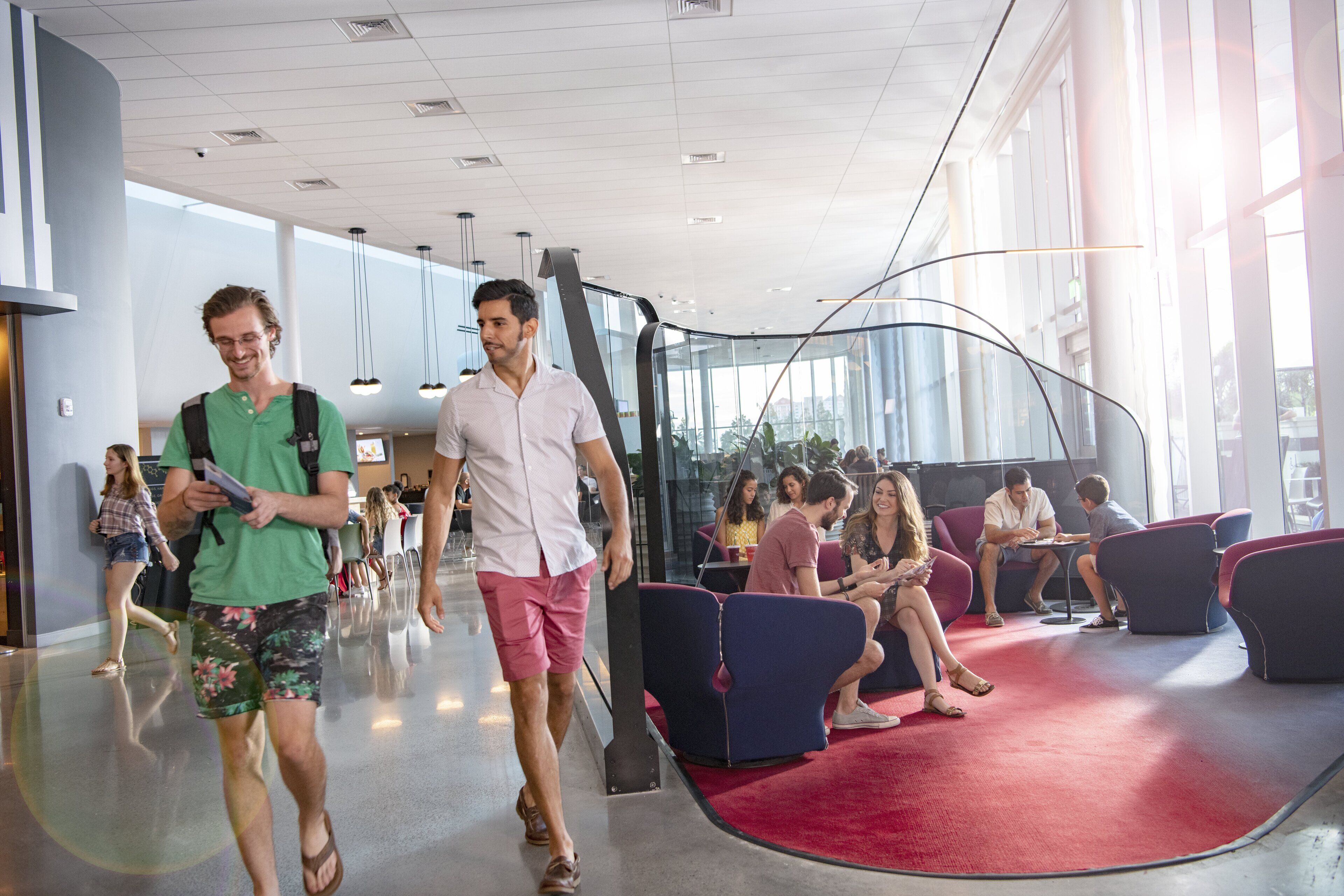 A modern indoor space with a curved glass wall, seating areas with red carpets, and people walking and sitting.