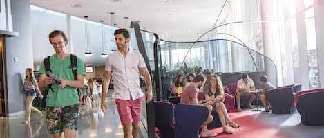 A modern indoor space with a curved glass wall, seating areas with red carpets, and people walking and sitting.