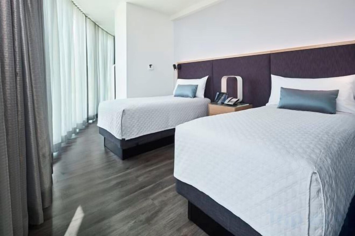 A hotel room with two beds, a nightstand, and a view of the outdoors.