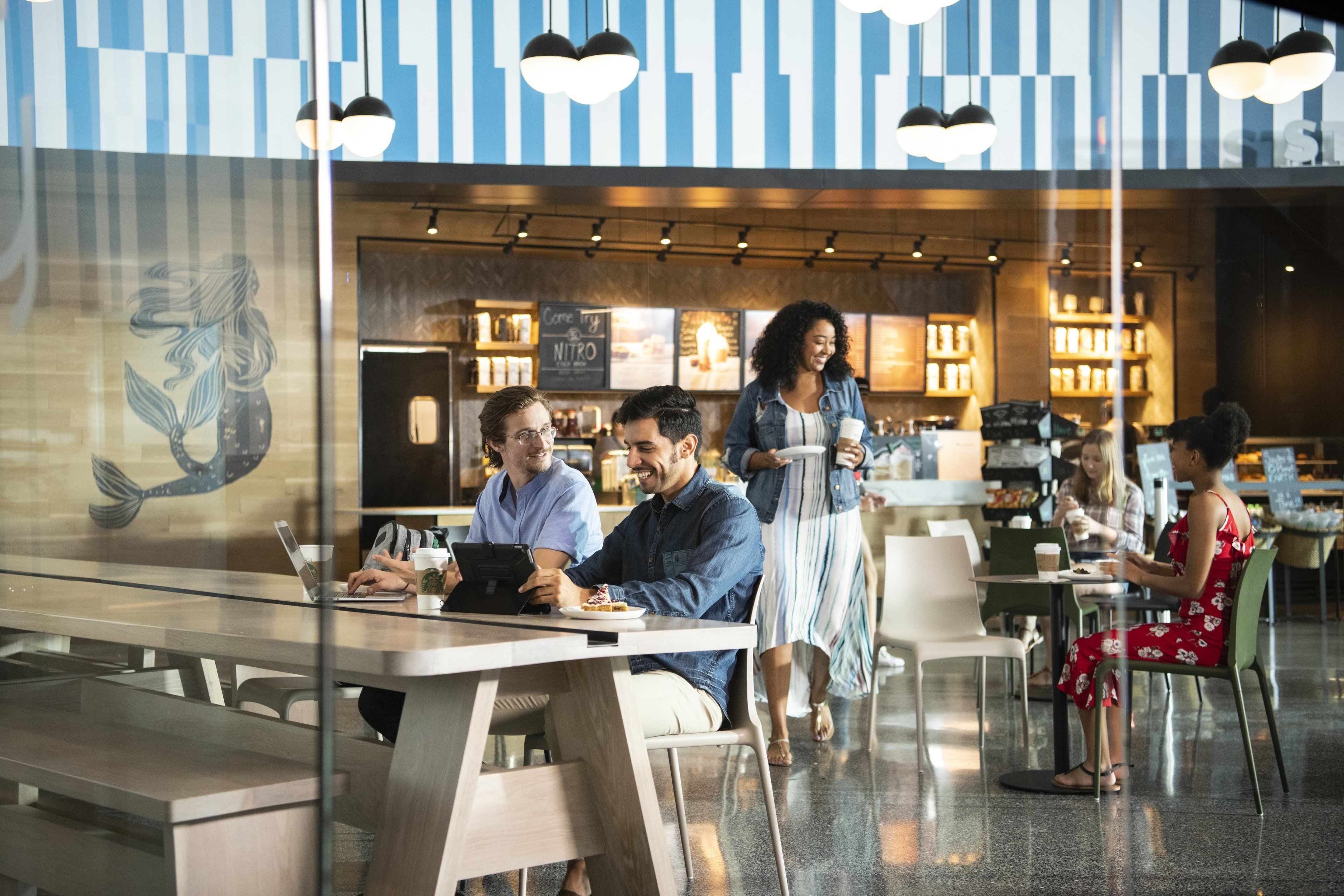 A modern cafe with a glass partition, people sitting at tables, and a menu board in the background.