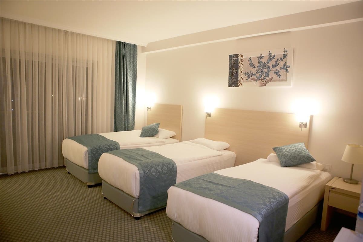 Habitación de hotel con dos camas, un ventanal grande con cortinas traslúcidas, una obra de arte en la pared y una mesita de noche con lámpara.