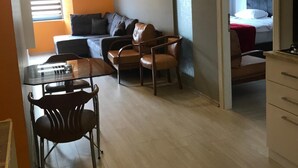 Una sala moderna con un sofá, dos sillones, una mesa de centro de vidrio y una mesa de comedor con sillas. Se ve una escalera al fondo que conduce a un piso superior.