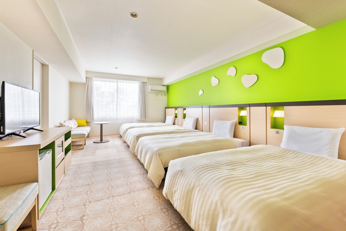 Habitación de hotel con tres camas, un escritorio con televisor, una mesita pequeña y una ventana con cortinas.
