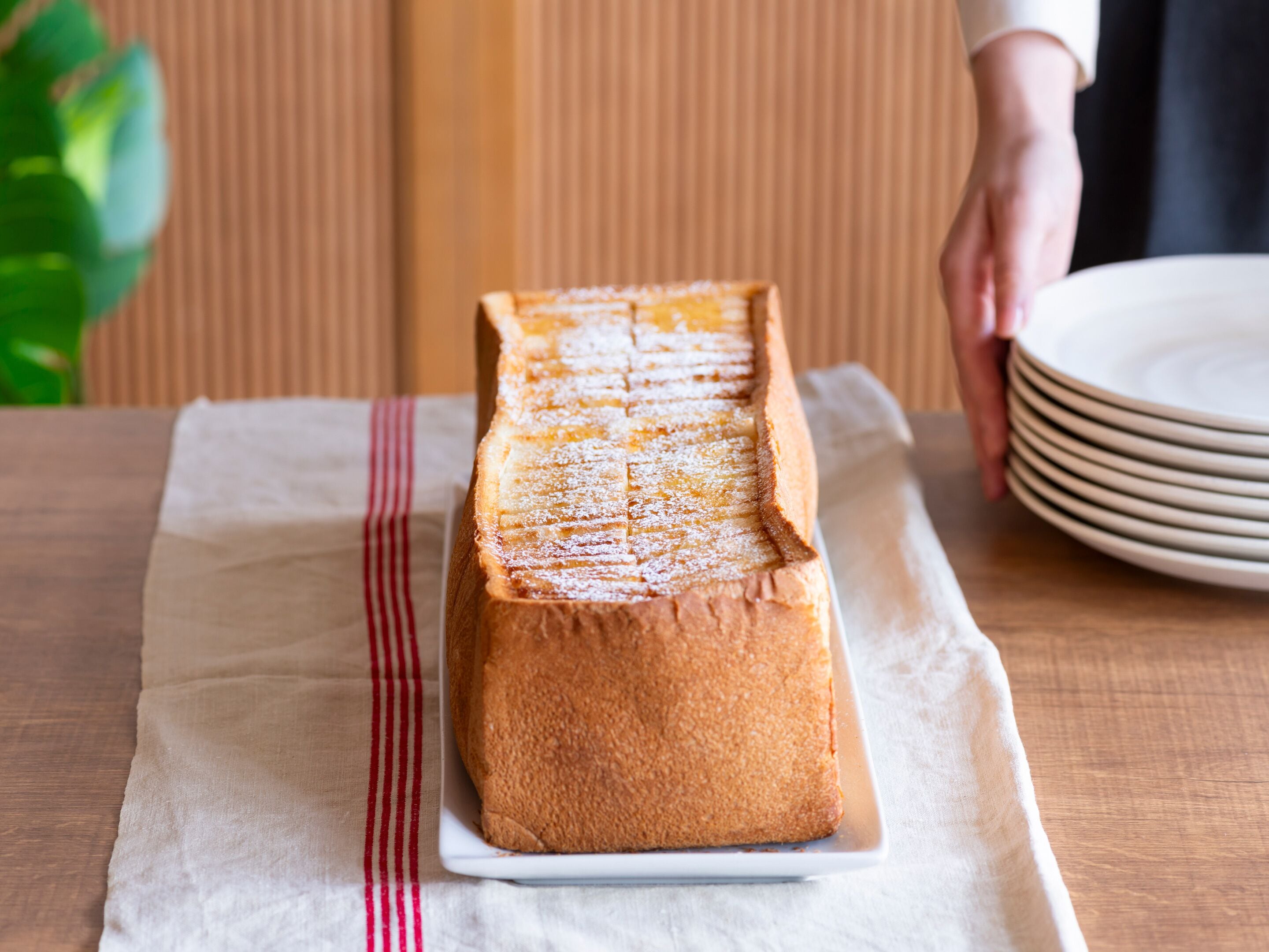 A loaf of bread on a white plate with a red stripe, placed on a white cloth on a wooden table.