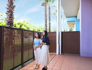 Dos personas de pie en un espacio exterior techado con piso de baldosas, cerca de una valla metálica y con palmeras al fondo.