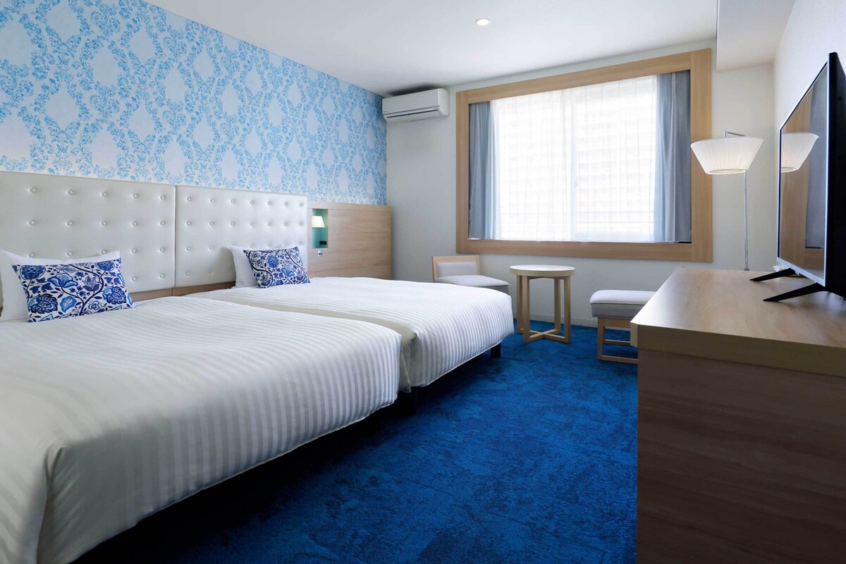 Habitación de hotel con dos camas, un escritorio, una silla, un televisor y una ventana con cortinas.