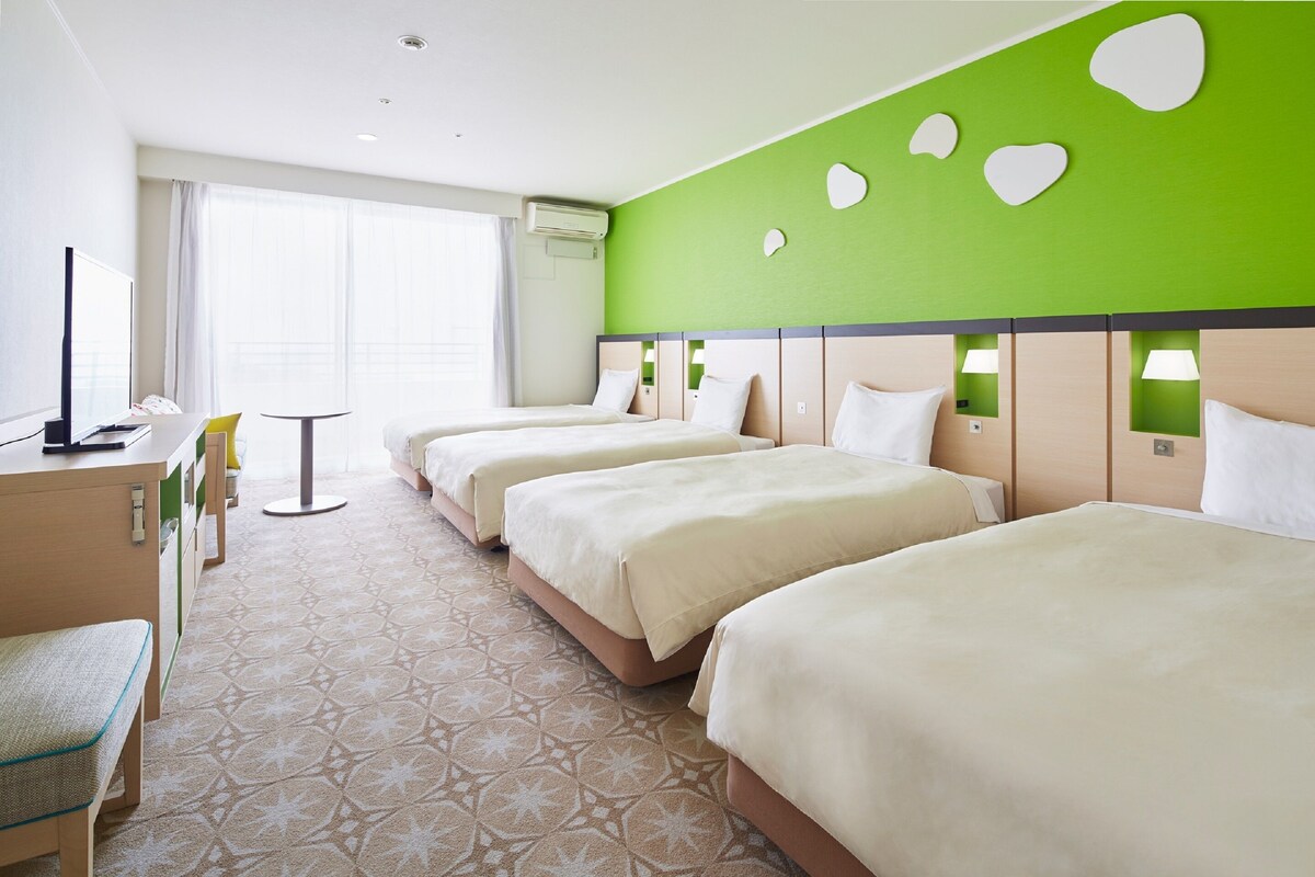 Habitación de hotel con tres camas, un escritorio, una silla y un amplio ventanal con cortinas.