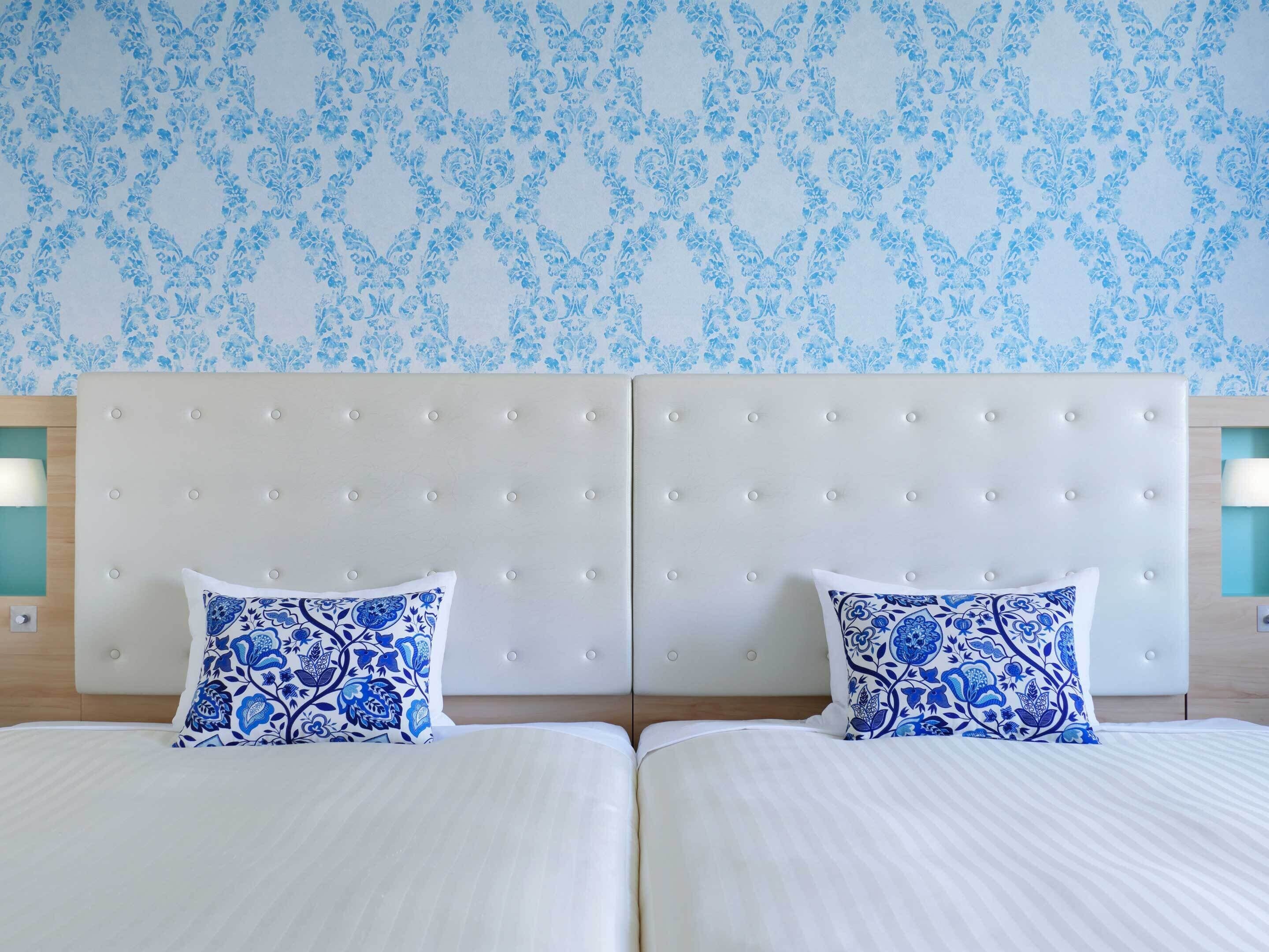 Una cama doble con cabecera blanca y dos almohadas decorativas, una mesita de noche de madera y una pared con estampado floral.