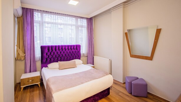 A bedroom with a large bed, a nightstand, a small stool, a window with curtains, and a mirror on the wall.