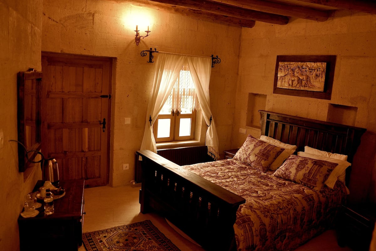 A bedroom with a wooden bed, a window with curtains, a wall-mounted light fixture, and a framed picture.