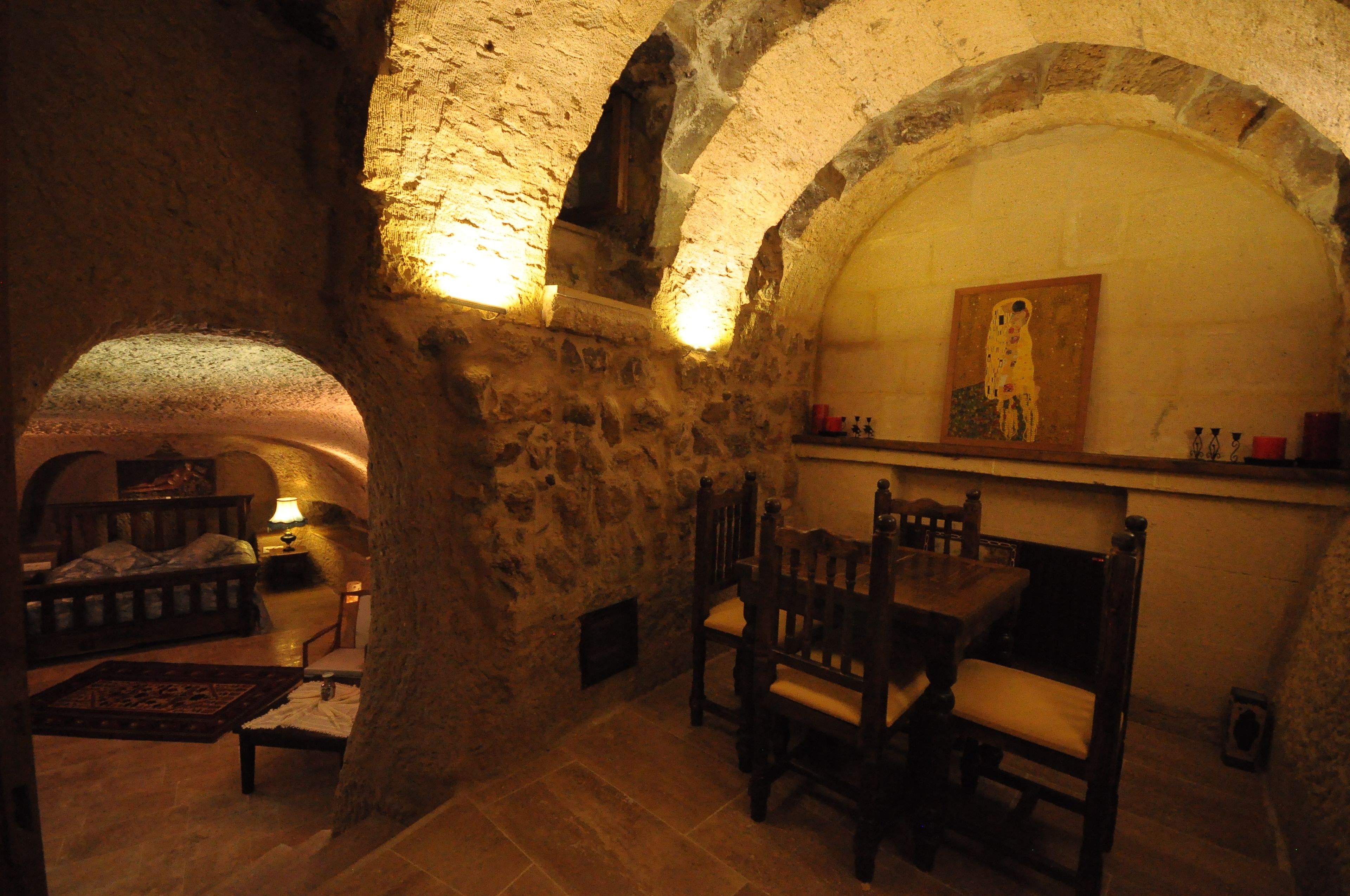A rustic stone interior with a bed, wooden chairs, and a painting on the wall.