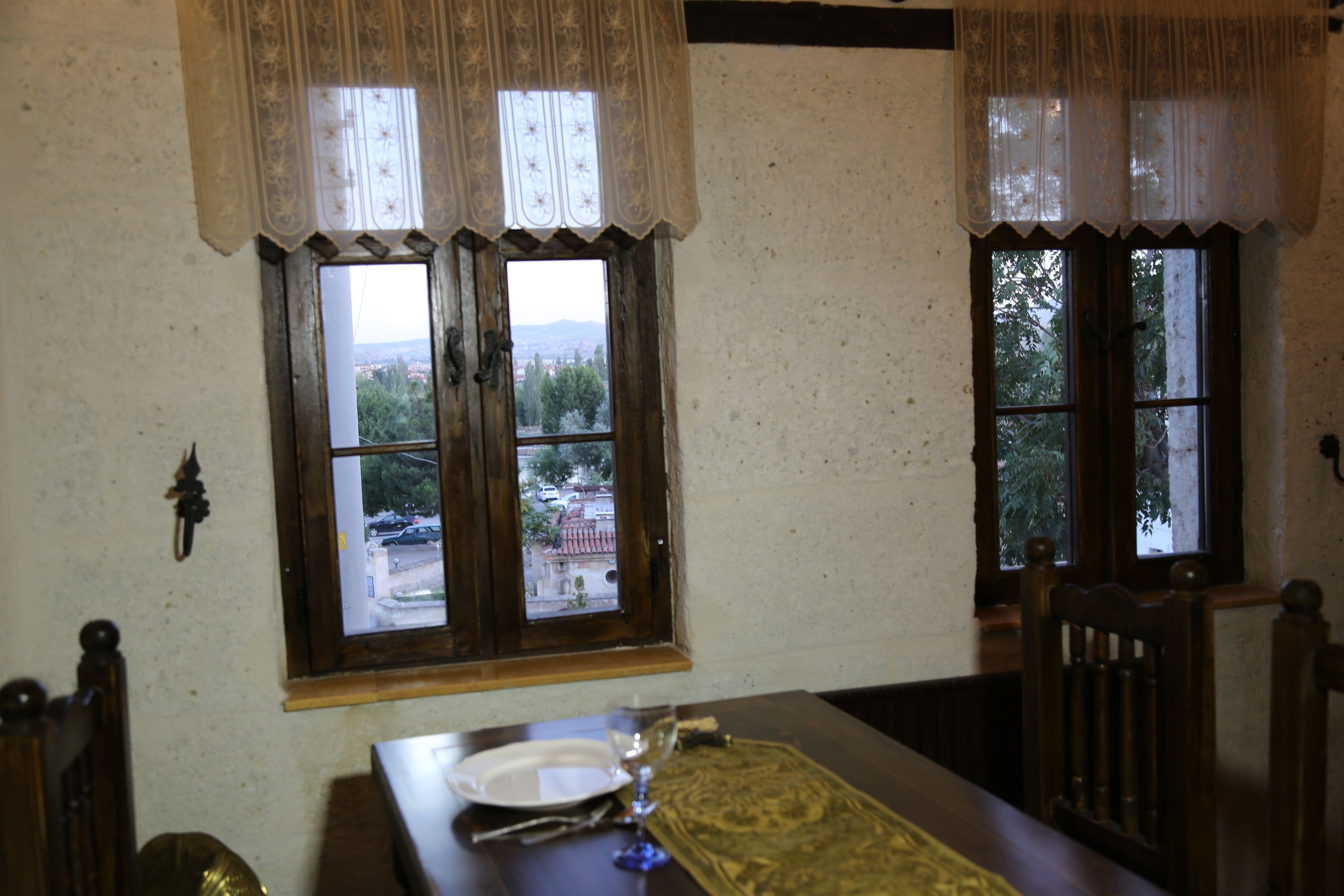 Un área de comedor con una mesa dispuesta para dos personas, dos ventanas con cortinas y vistas a un paisaje urbano.