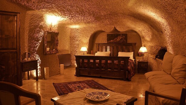 A cozy, rustic room with a vaulted ceiling, a wooden bed, a sofa, and a dining table with a plate. - Hanımkız Konagi (Avanos)
