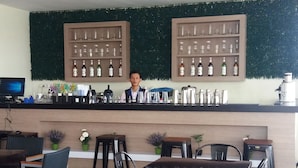 Una zona de bar con un camarero, estantes llenos de botellas y utensilios de bar. Se ven sillas y mesas en primer plano.