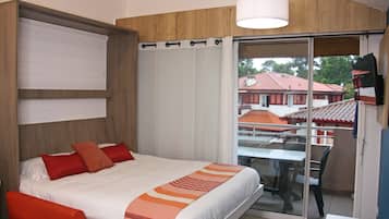 A hotel room with a bed, a table, and a chair. There is a balcony with a view of buildings.