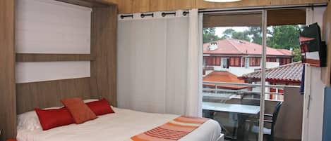 A hotel room with a bed, a table, and a chair. There is a balcony with a view of buildings.