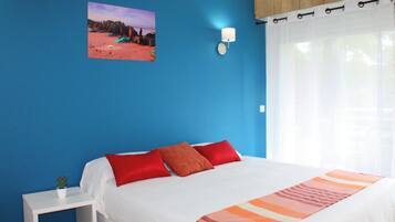 Une chambre à coucher avec un grand lit, une table de chevet, une lampe fixée au mur et une fenêtre ornée de rideaux légers.