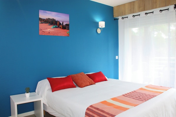 A bedroom with a large bed, a nightstand, a wall-mounted lamp, and a window with sheer curtains. - Résidence du Parc (Soorts-Hossegor)