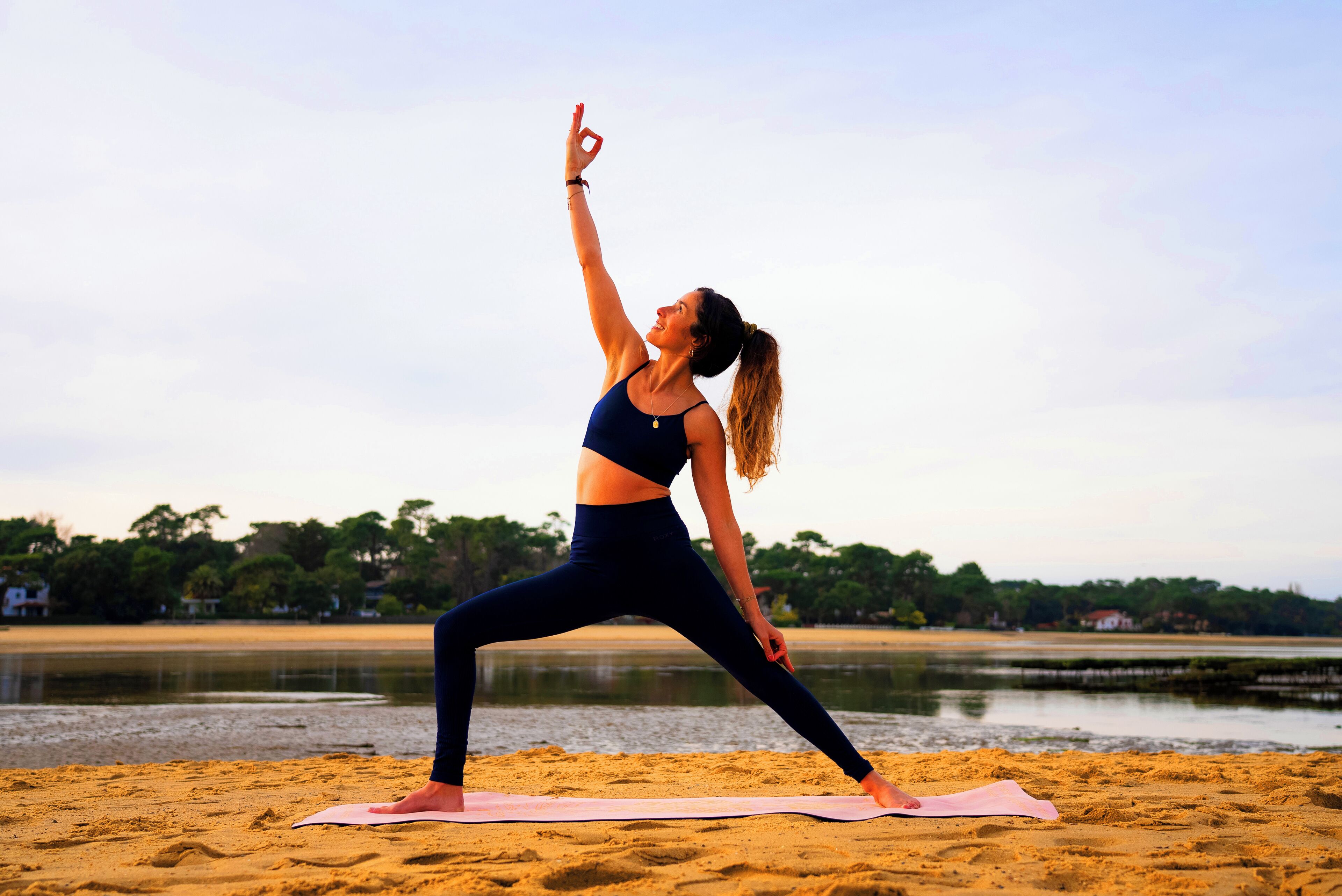 A person performing a yoga pose on a beach with a calm sea and trees in the background.