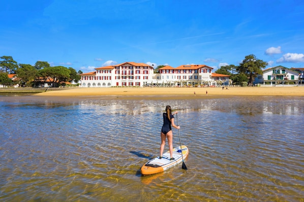 A woman paddleboarding on a calm lake with a large building in the background. - Résidence du Parc (Soorts-Hossegor)