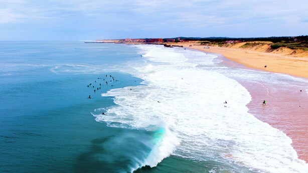 Una playa con olas rompiendo en la orilla, arena y una costa distante con edificios.